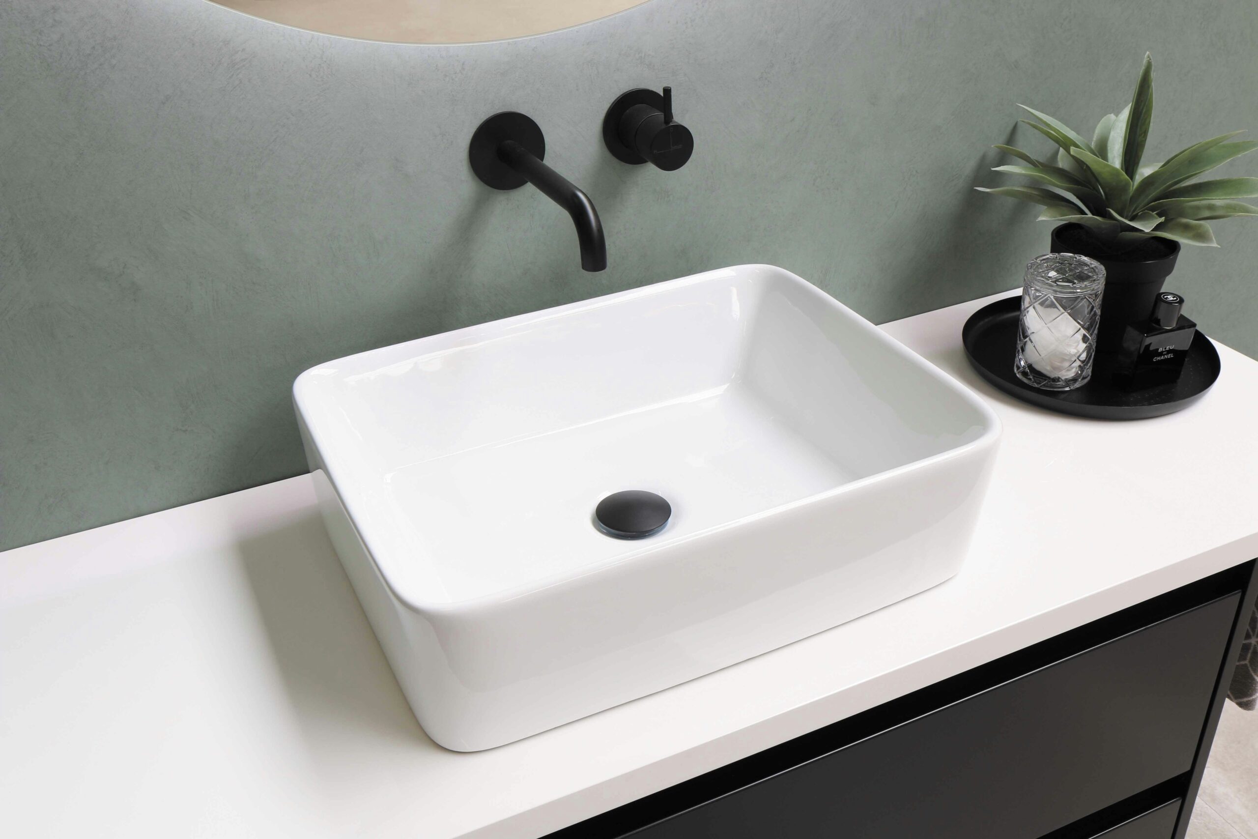 Lavabo rectangular blanco sobre encimera blanca, con grifería negra de pared y fondo verde suave.