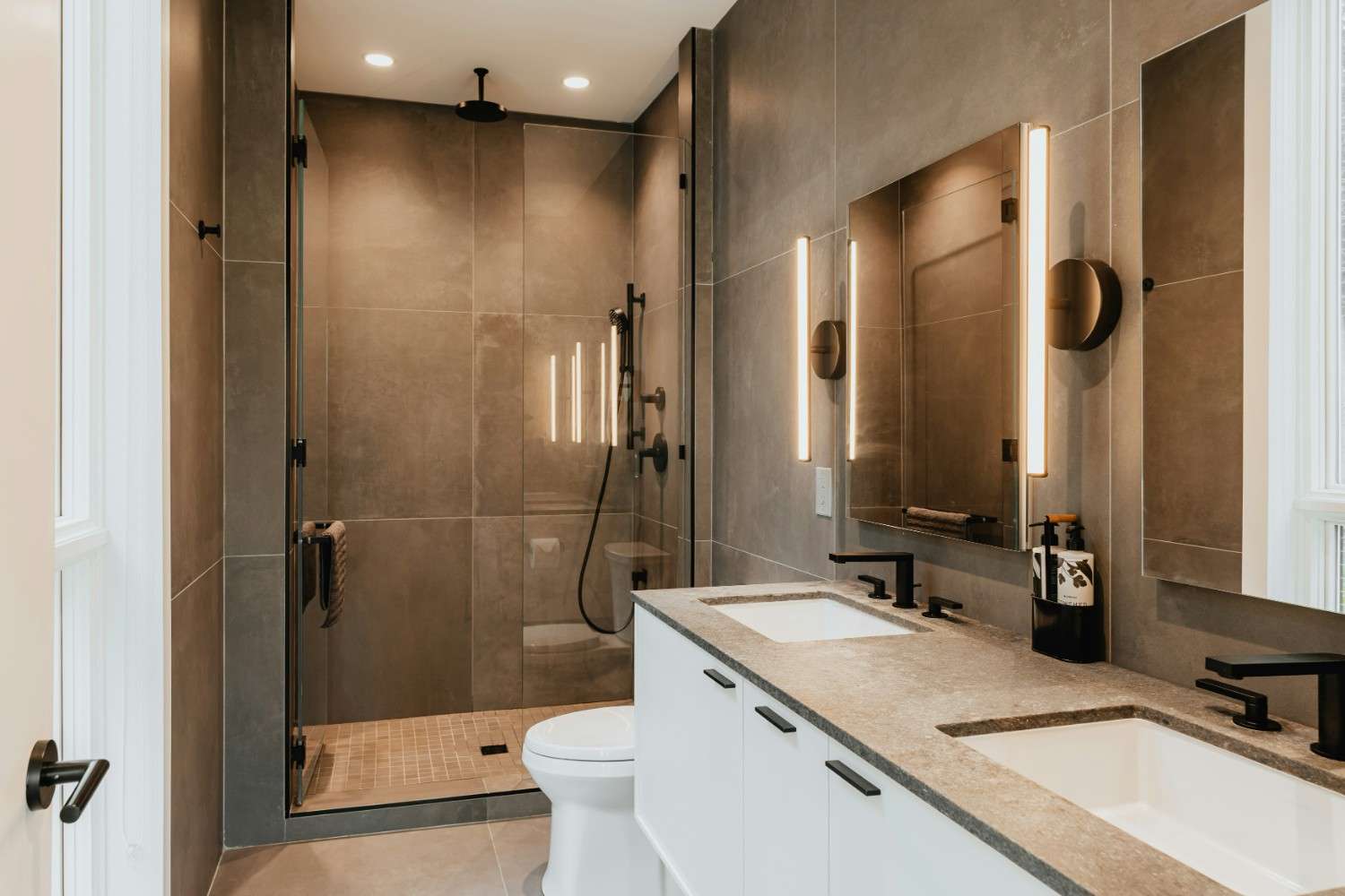 Baño con mueble blanco de doble lavabo, grifería negra, espejos con iluminación lateral y ducha con mampara de cristal.