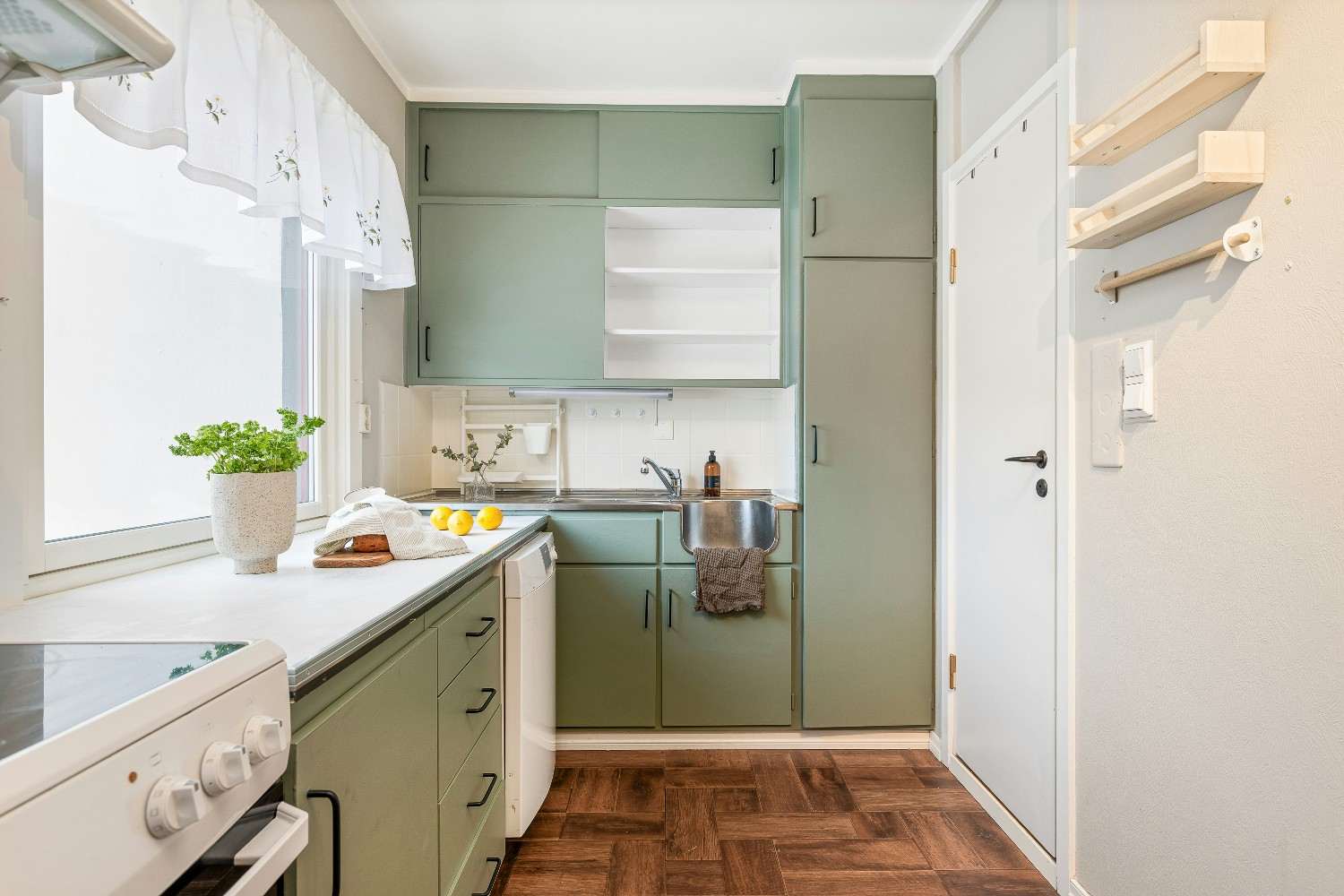 Cocina pequeña con muebles blancos, pared verde y suelo de madera.