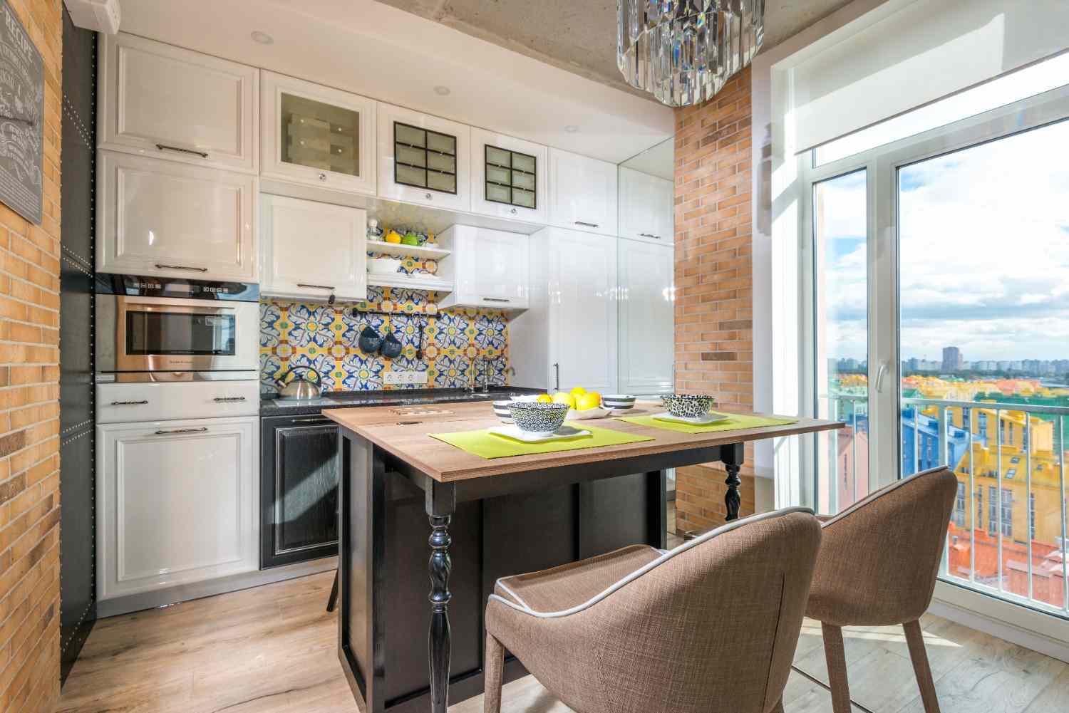 Cocina pequeña con muebles blancos, azulejos de colores y mesa junto al ventanal.