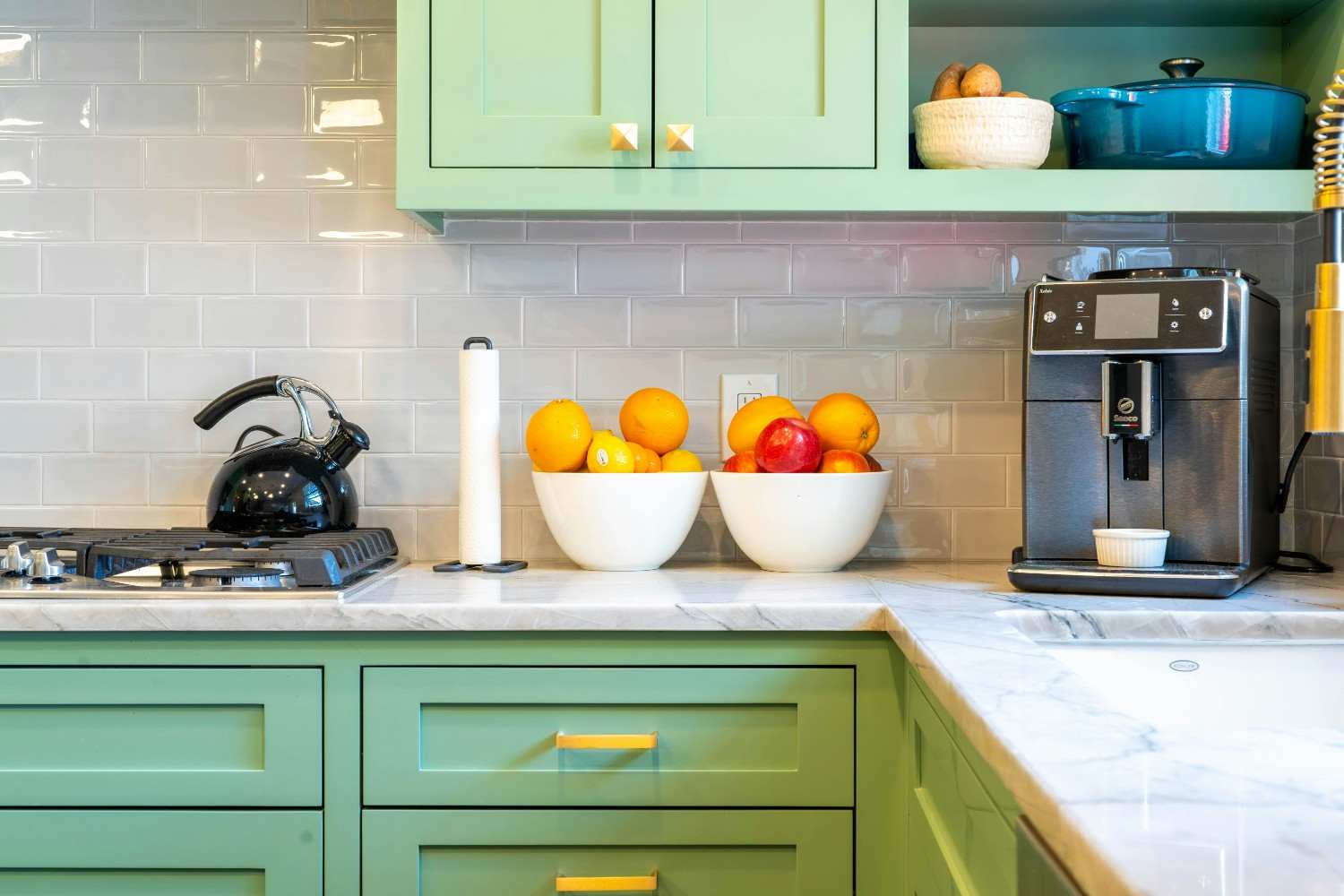 Cocina con muebles verdes, tiradores dorados, encimera clara y cuencos de fruta junto a una cafetera.