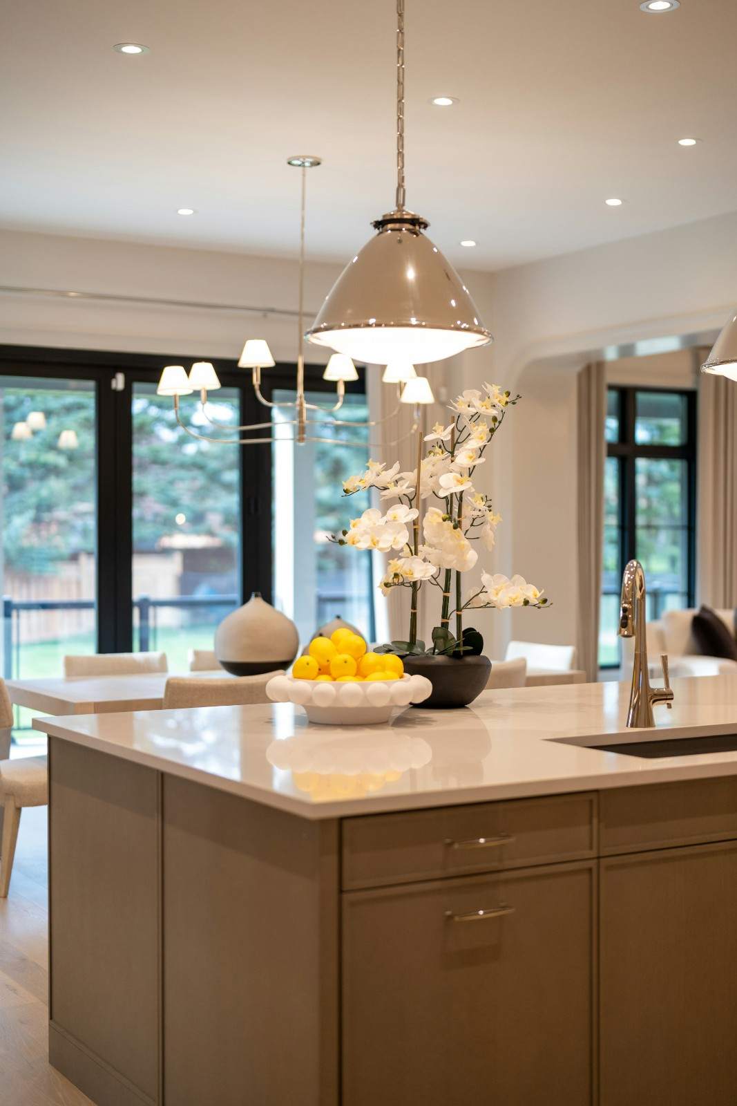 Isla de cocina con muebles de madera, encimera clara, una orquídea blanca y un frutero con limones bajo una lámpara colgante.