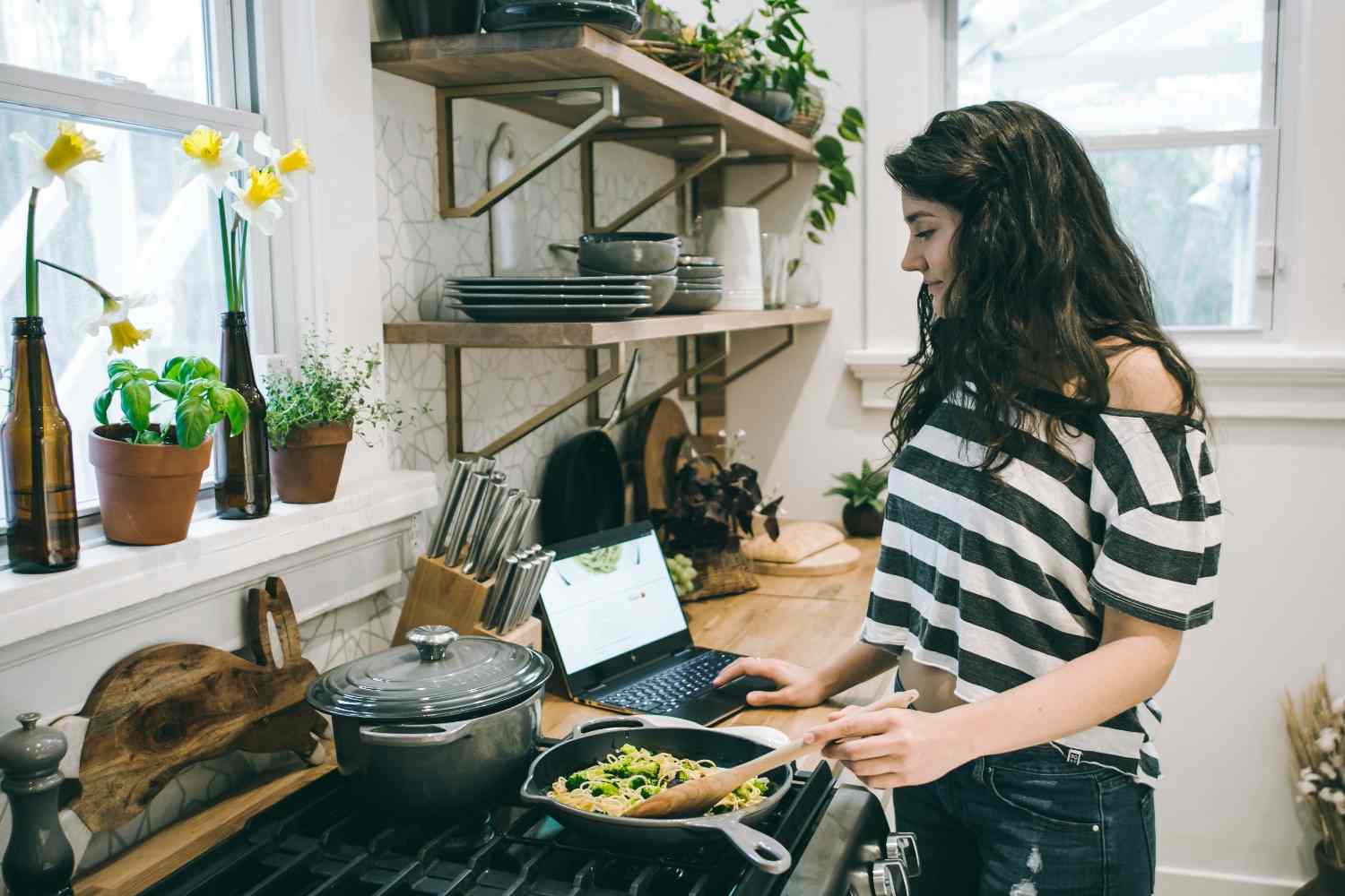 Mujer cocinando en una cocina con estantes abiertos y plantas junto a la ventana.