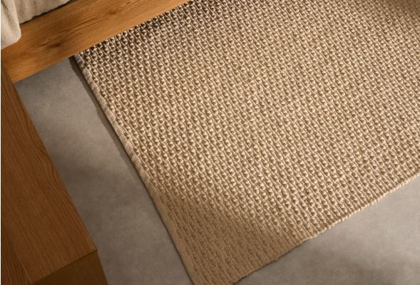 Detalle del tejido de una alfombra de tono crudo con pequeños relieves junto a una cama de madera