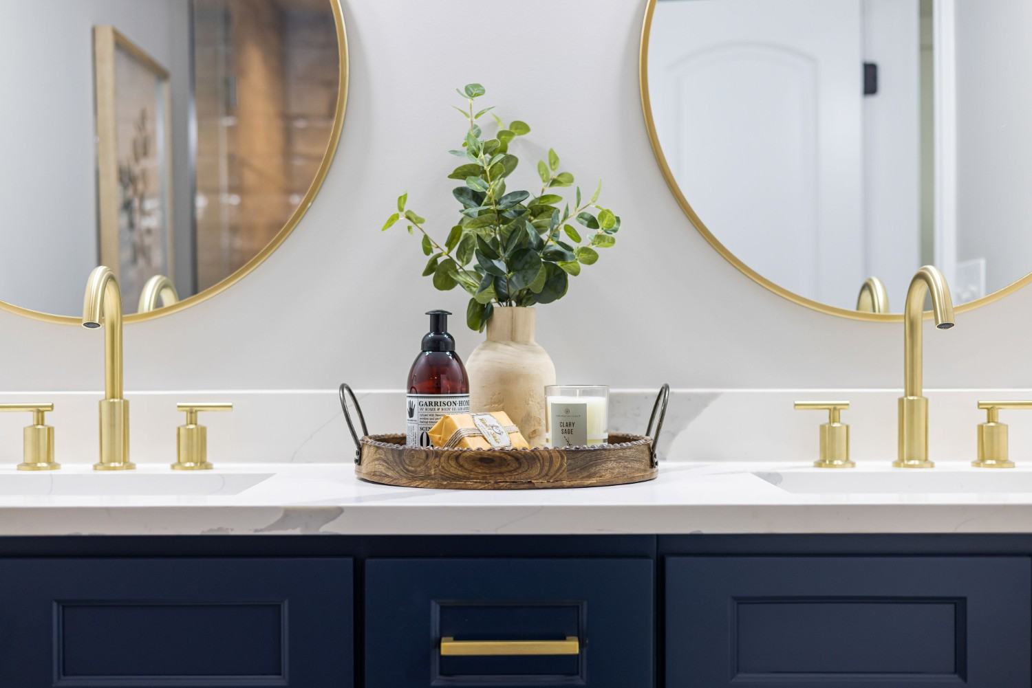 Mueble azul marino con doble lavabo blanco, grifería dorada y dos espejos redondos con marcos dorados.