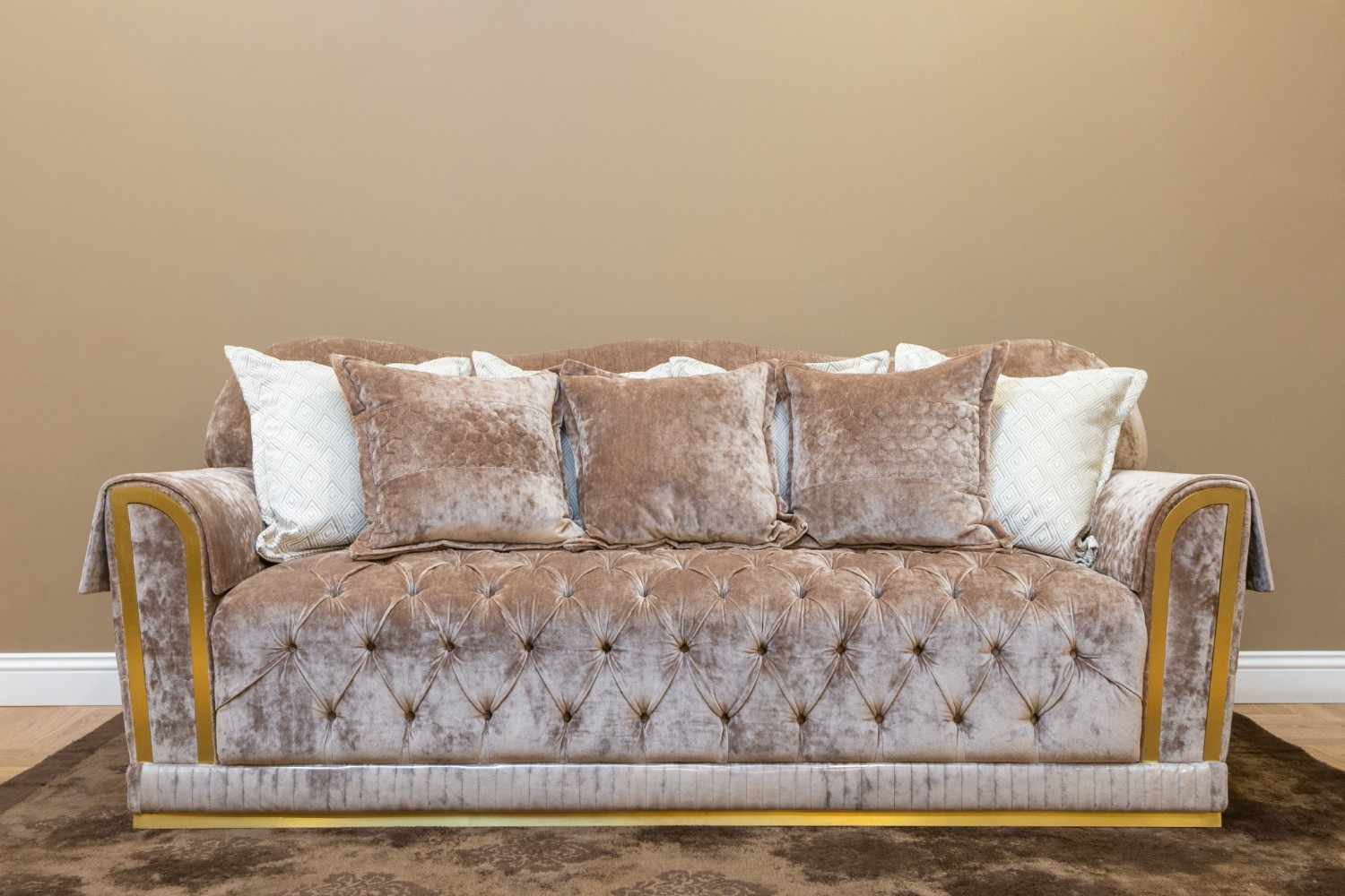 Sofá recto de terciopelo beige con capitoné, cojines y detalles dorados en los brazos y la base.
