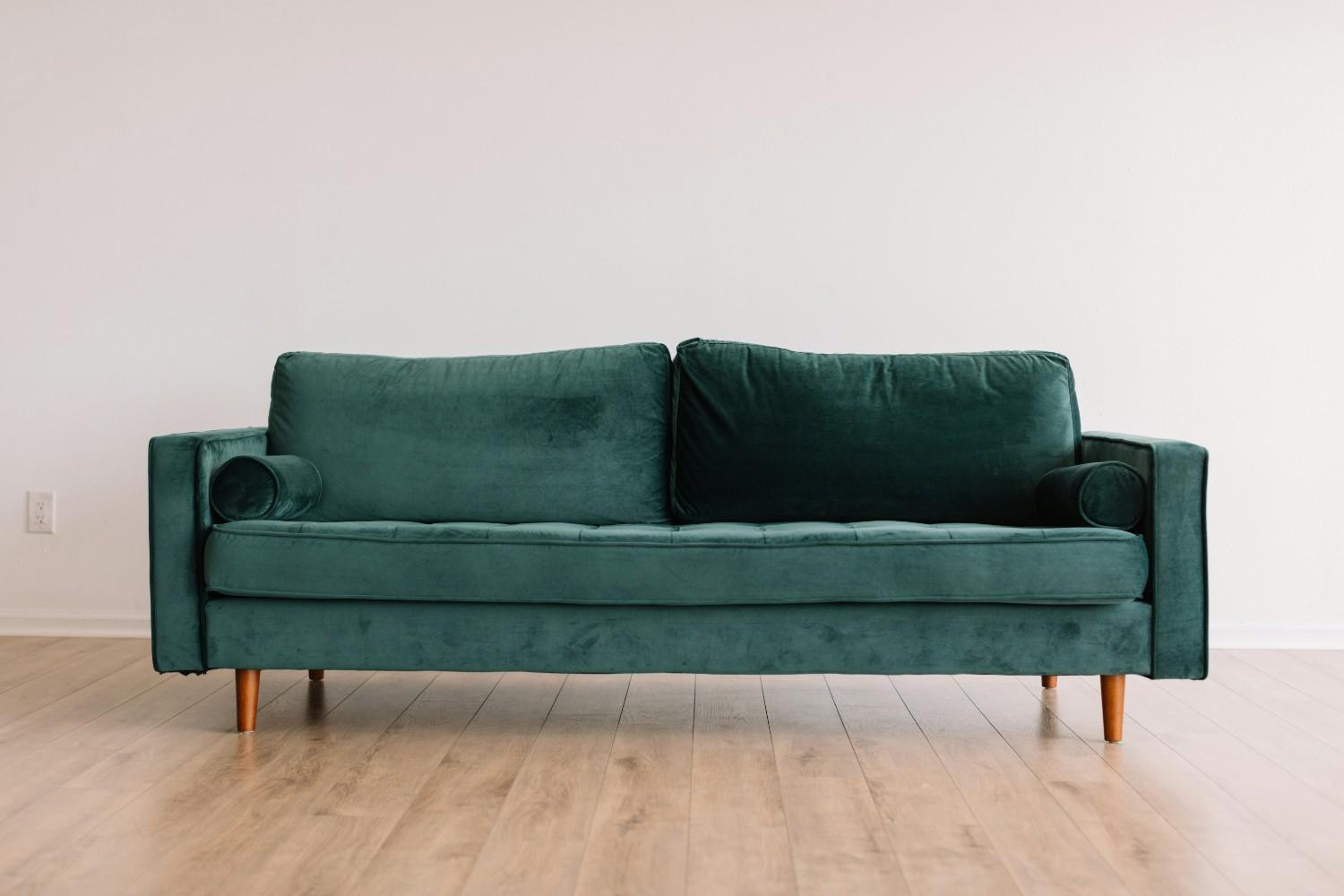 CÓMO ELEGIR UN SOFÁ QUE ENCAJE EN TU SALÓN Y EN TU VIDA Sofá verde con cojines cilíndricos y patas de madera sobre un suelo de madera clara.